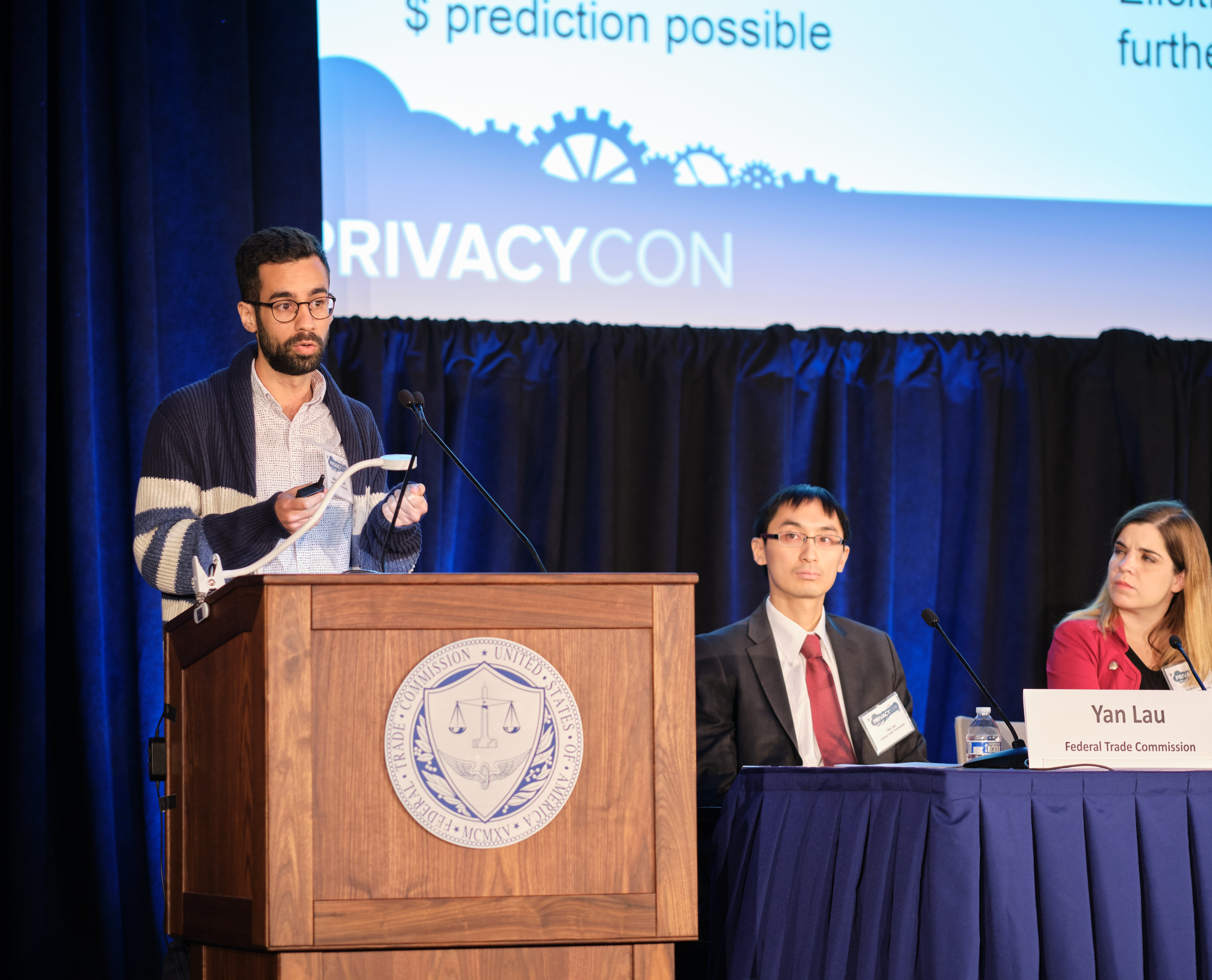 Man standing at podium at a conference with two people listening from a long table on stage